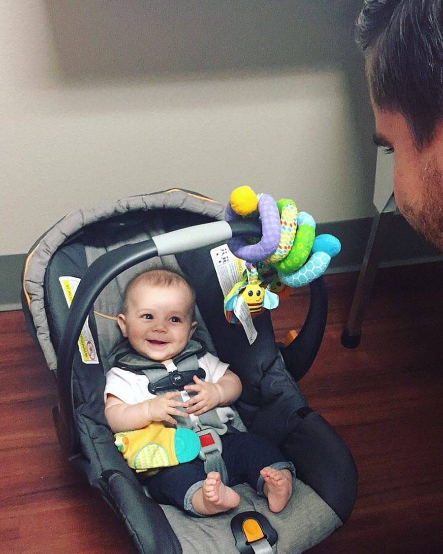 A happy, smiling baby in a car seat being greeted at The Roots
