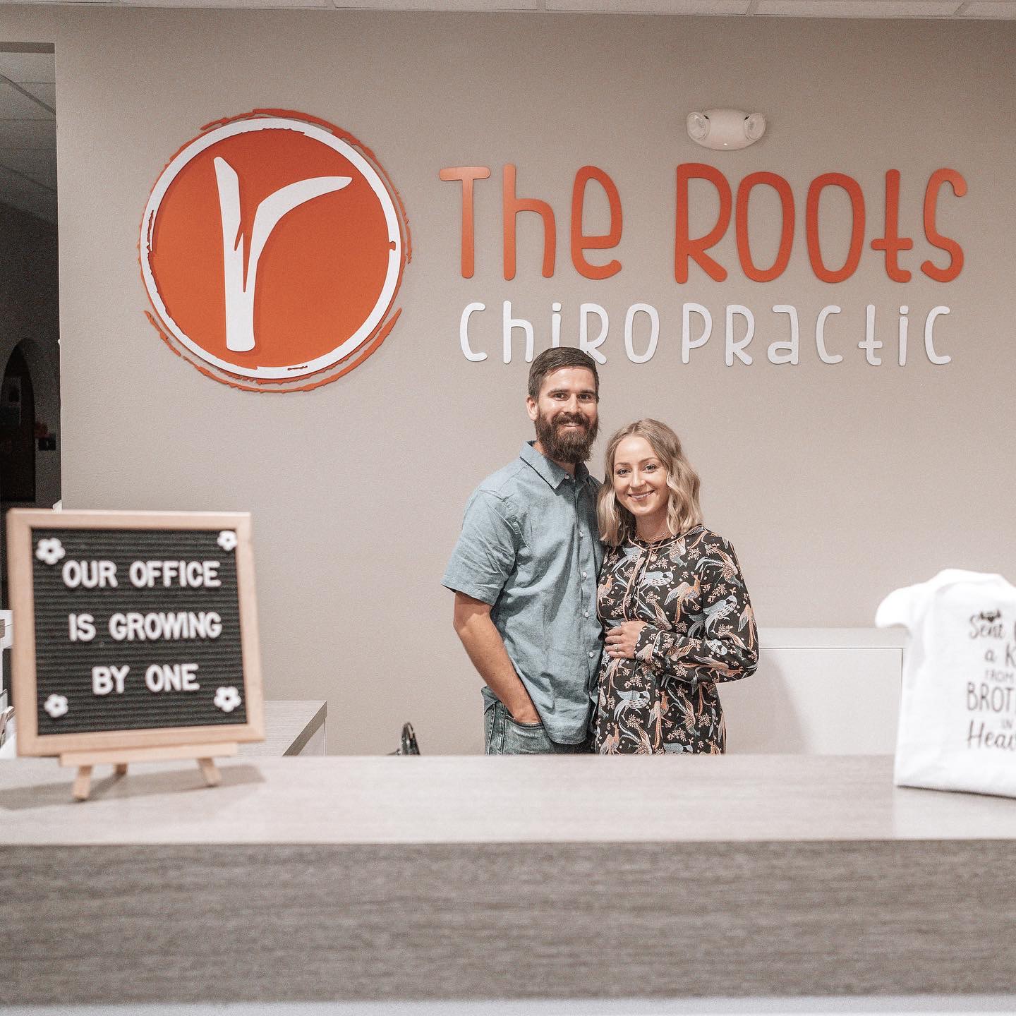 Dr. Logan and Dr. Laura Swaim, co-directors, smiling together at the front desk beneath the orange Roots Chiropractic logo
